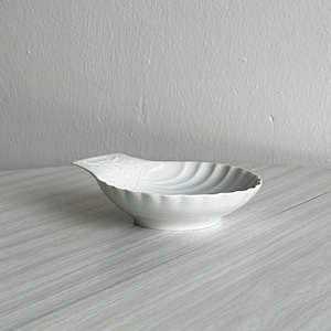 May include: A white ceramic dish shaped like a scallop shell with a fluted interior and a small handle on one side. The bowl features a smooth, glossy finish and a scalloped edge, resting on a circular base with a shallow, wide basin design.