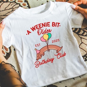 Könnte beinhalten: Weißes T-Shirt mit einer Dackel-Grafik, die eine Partyhütchen trägt und Luftballons hält. Roter Text lautet "A Weenie Bit Older Birthday Club EST 2025". Das Shirt wird von Händen gehalten.