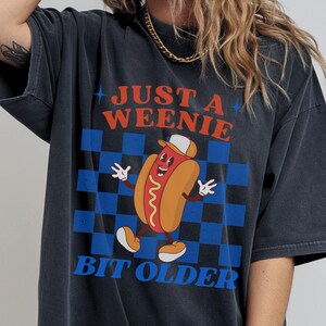 May include: Dark gray t-shirt featuring a cartoon hot dog wearing a cap and smiling. The shirt has the text "JUST A WEENIE BIT OLDER" in red and blue, set against a blue and black checkered background.