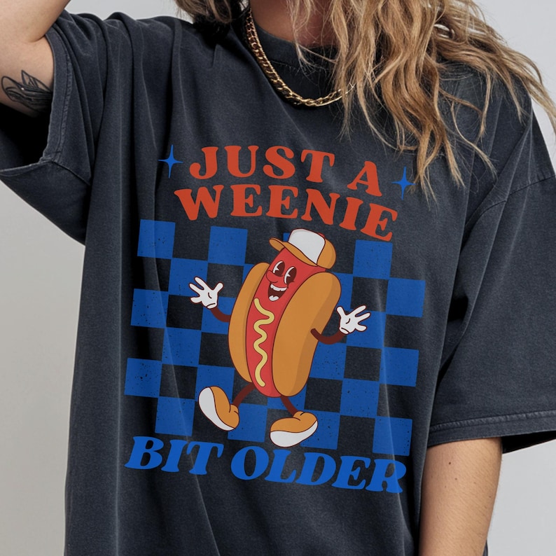 May include: Dark gray t-shirt featuring a cartoon hot dog wearing a cap and smiling. The shirt has the text "JUST A WEENIE BIT OLDER" in red and blue, set against a blue and black checkered background.