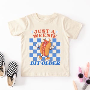 May include: Cream-colored t-shirt with the text "JUST A WEENIE BIT OLDER" and a cartoon hot dog character. The hot dog wears a hat and is set against a blue and white checkered background. Black and white checkered sneakers are visible.