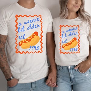 Könnte beinhalten: Cremefarbene T-Shirts mit einem Hotdog-Motiv in einem wellenförmigen orangefarbenen Rahmen. Der Text "a weenie bit older est. 1995" steht in blauer Schrift. Zwei Personen tragen die Shirts und halten Händchen.