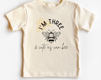 May include: A cream colored t-shirt with a black bee and honeycomb graphic. The text "I'm Three & cute as can bee" is printed on the shirt.