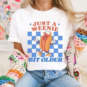 May include: White t-shirt with a cartoon hot dog graphic. The hot dog is wearing a hat and has a smiling face. The shirt has the text "JUST A WEENIE" above the graphic and "BIT OLDER" below it, all in a retro font.
