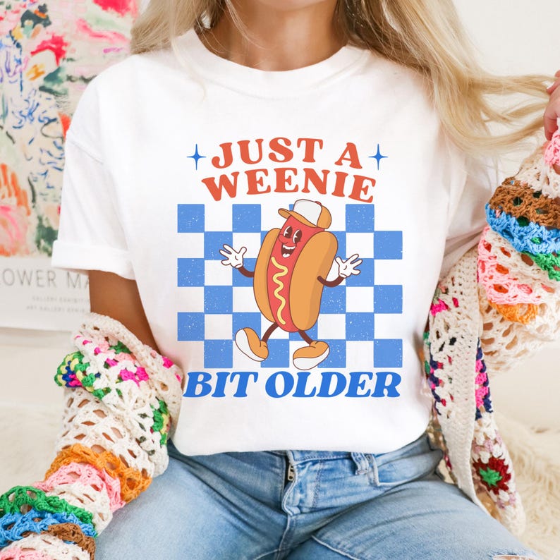 May include: White t-shirt with a cartoon hot dog graphic. The hot dog is wearing a hat and has a smiling face. The shirt has the text "JUST A WEENIE" above the graphic and "BIT OLDER" below it, all in a retro font.