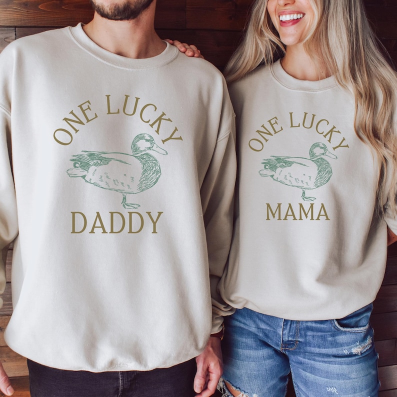 以下が含まれることがあります： 緑色のアヒルのイラストと「One Lucky Daddy」と「One Lucky Mama」の文字が書かれた、クリーム色のスウェットシャツ2枚。