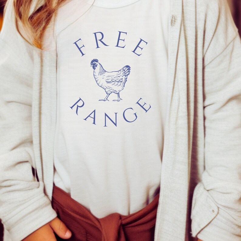 May include: Cream-colored t-shirt with the words "FREE RANGE" in navy blue arched above and below a detailed illustration of a chicken. The shirt is worn with a light gray cardigan and a rust-colored bottom.