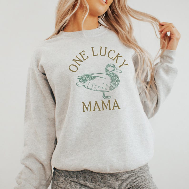 以下が含まれることがあります： 茶色の文字で「One Lucky Mama」と書いてある灰色のスウェットシャツ。緑色のアヒルのイラストが描かれている。