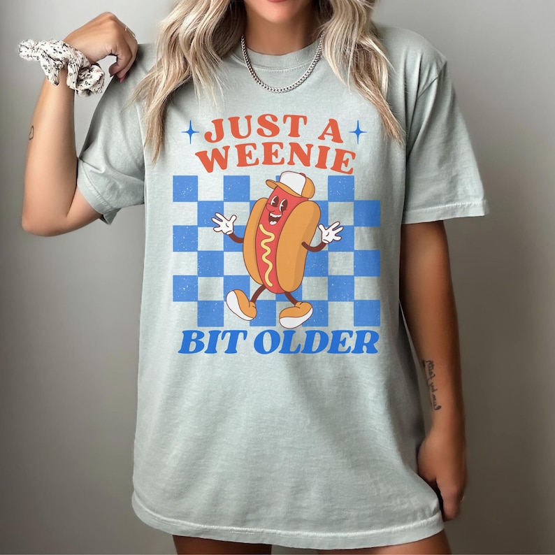 May include: A light green t-shirt with the text "JUST A WEENIE BIT OLDER" above and below a cartoon hot dog character. The background is a blue and white checkered pattern.