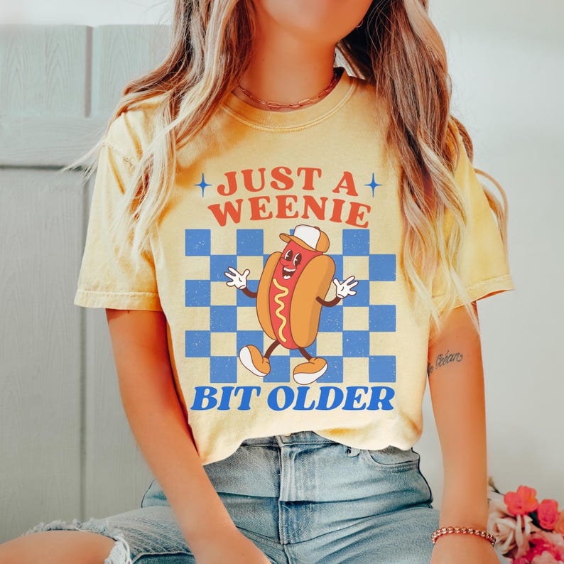 May include: Yellow t-shirt with the text "JUST A WEENIE BIT OLDER" and a cartoon hot dog character wearing a hat. The background features a blue and white checkered pattern.