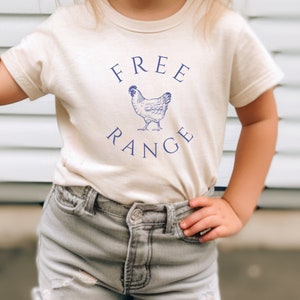 May include: A cream-colored t-shirt featuring the words "FREE RANGE" in navy blue, with a detailed illustration of a chicken in the center. The shirt is short-sleeved and appears to be made of soft material.