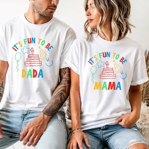 May include: White t-shirts with the text "It's Fun to Be Dada" and "It's Fun to Be Mama" in colorful lettering. The shirts feature a birthday cake, party hat, and balloon graphic. The shirts are worn by a man and a woman.
