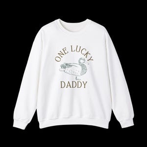 以下が含まれることがあります： 白地に緑色のアヒルのイラストと、茶色の文字で「One Lucky Daddy」と書かれたスウェットシャツです。