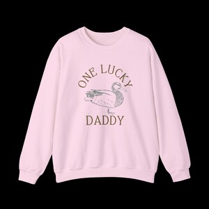 以下が含まれることがあります： 「One Lucky Daddy」という文字とアヒルの絵が描かれたピンク色のスウェットシャツです。