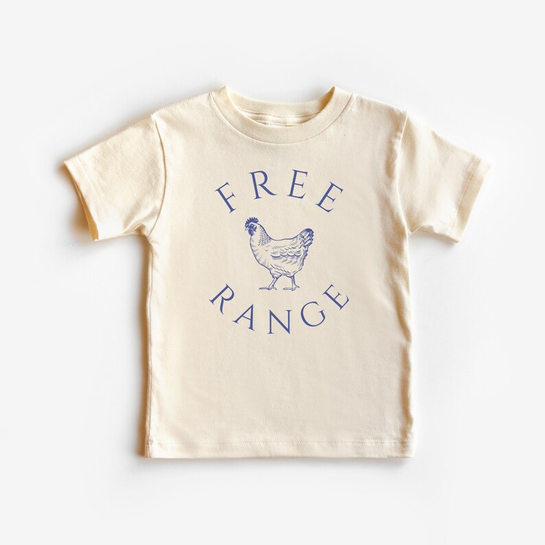 May include: Cream-colored short-sleeved t-shirt with the words "FREE RANGE" in navy blue arched above and below a navy blue illustration of a chicken. The shirt is made of soft material.