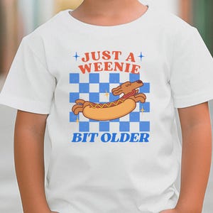 Op de afbeelding: Wit T-shirt met een cartoon hotdog ontwerp. Het ontwerp bevat de tekst "JUST A WEENIE" boven een cartoon hotdog en de tekst "BIT OLDER" eronder. De achtergrond is een blauw-wit geblokt patroon.
