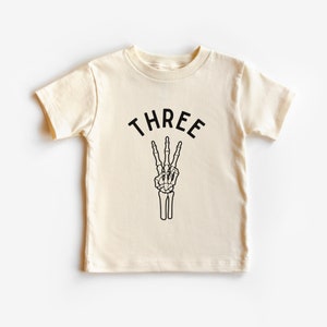 Puede incluir: Camiseta crema para niño con la palabra "THREE" sobre una mano de esqueleto negra haciendo el signo de la paz. La camiseta de manga corta es de material suave y es adecuada para el tercer cumpleaños de un niño.