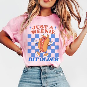 May include: Pink t-shirt with a graphic of a cartoon hot dog wearing a hat, set against a blue and white checkered background. The text reads "JUST A WEENIE BIT OLDER" in orange and blue.
