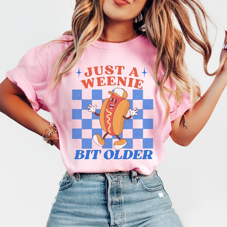 May include: Pink t-shirt with a graphic of a cartoon hot dog wearing a hat, set against a blue and white checkered background. The text reads "JUST A WEENIE BIT OLDER" in orange and blue.