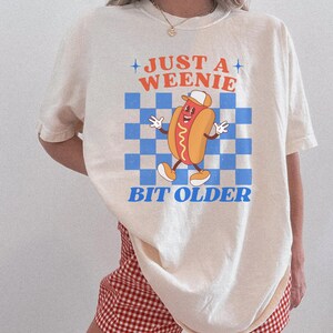 May include: Cream-colored t-shirt featuring a cartoon hot dog character wearing a hat, set against a blue and white checkered background. The text "JUST A WEENIE BIT OLDER" is printed above and below the graphic.