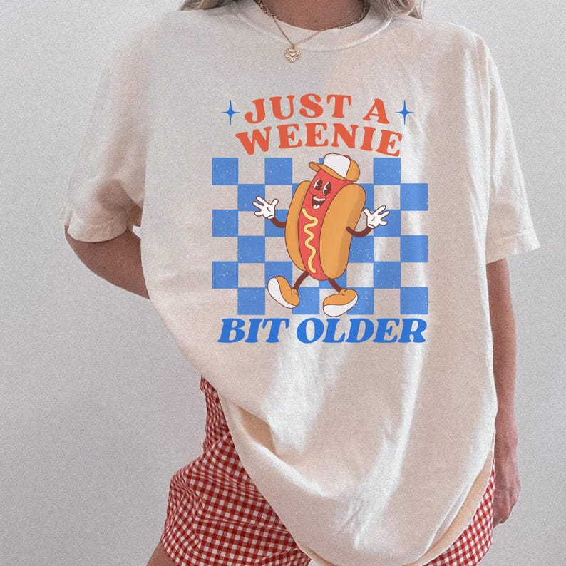 May include: Cream-colored t-shirt featuring a cartoon hot dog character wearing a hat, set against a blue and white checkered background. The text "JUST A WEENIE BIT OLDER" is printed above and below the graphic.