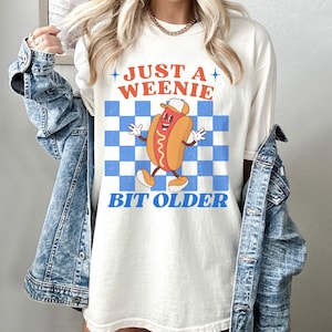May include: Cream-colored t-shirt featuring a cartoon hot dog character with a hat, arms, and legs, set against a blue and white checkered background. The text "JUST A WEENIE" and "BIT OLDER" are printed above and below the graphic.