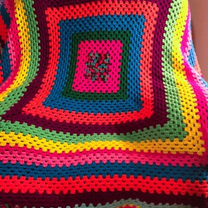 May include: A vibrant, handmade crochet blanket with a square pattern. The blanket features concentric squares of bright colors, including pink, blue, green, yellow, and purple. The blanket is likely made of soft yarn and is suitable for use as a throw or decorative piece.