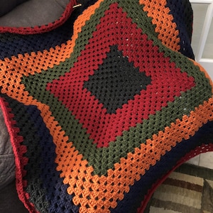 May include: A handmade crochet blanket with a geometric pattern. The blanket features a central dark green square, surrounded by red, olive green, orange, and navy blue bands. The edges are finished with a red border.
