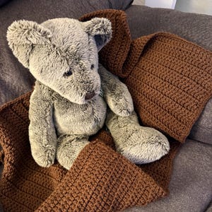 May include: A plush gray teddy bear is nestled in a brown crocheted blanket. The bear has a soft, furry texture and is positioned on a gray sofa. The blanket is a warm, earthy tone, creating a cozy scene.