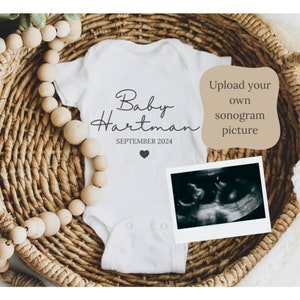 May include: White onesie with the text "Baby Hartman" and "SEPTEMBER 2024" printed on it. A small heart is printed below the date. A black and white sonogram image is in a white frame next to the onesie. The onesie is in a woven basket with wooden beads.