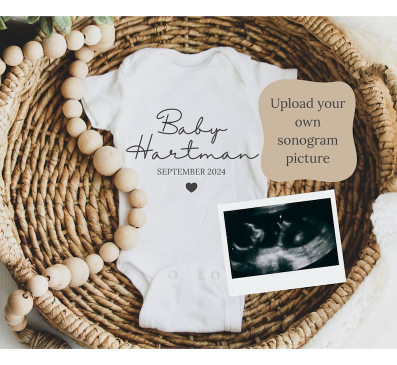 May include: White onesie with the text "Baby Hartman" and "SEPTEMBER 2024" printed on it. A small heart is printed below the date. A black and white sonogram image is in a white frame next to the onesie. The onesie is in a woven basket with wooden beads.