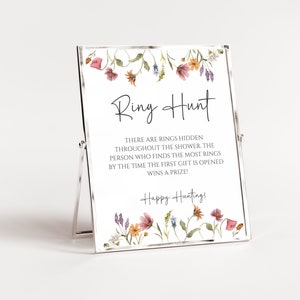 May include: A white sign with a floral border and the text "Ring Hunt" in black. The sign says "There are rings hidden throughout the shower. The person who finds the most rings by the time the first gift is opened wins a prize! Happy Hunting!"