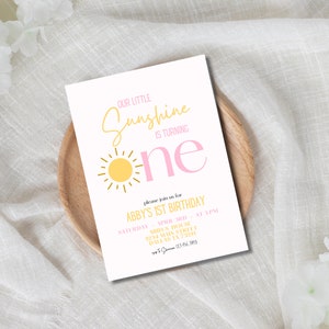K&ouml;nnte beinhalten: Eine rosafarbene und gelbe Geburtstagseinladung mit einer Sonnendarstellung und dem Text "Our Little Sunshine is Turning One". Die Einladung enth&auml;lt die Details f&uuml;r Abbys 1. Geburtstagsparty, einschlie&szlig;lich Datum, Uhrzeit und Ort.