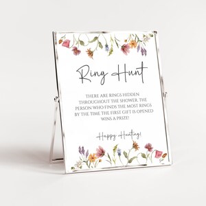 May include: A white sign with a floral border and the text "Ring Hunt" in black. The sign says "There are rings hidden throughout the shower. The person who finds the most rings by the time the first gift is opened wins a prize! Happy Hunting!"