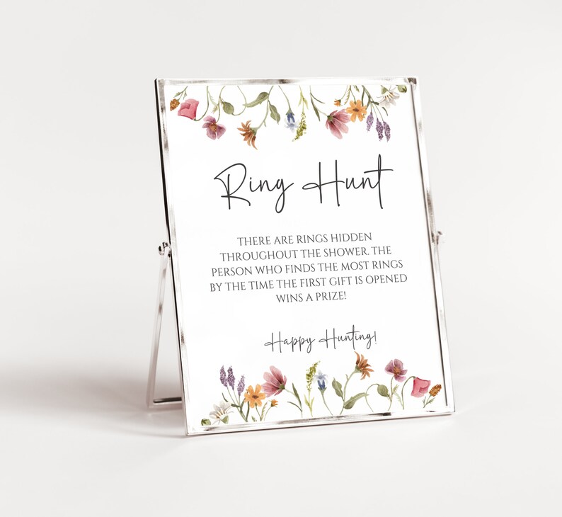 May include: A white sign with a floral border and the text "Ring Hunt" in black. The sign says "There are rings hidden throughout the shower. The person who finds the most rings by the time the first gift is opened wins a prize! Happy Hunting!"