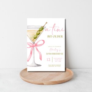 May include: A white invitation card with a pink ribbon and a martini glass with olives. The text on the card reads "a tini bit older" and "Harley's 30th Birthday". The invitation includes the date, time, and location of the party.