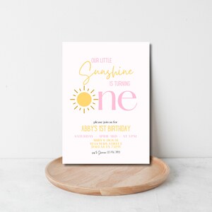 K&ouml;nnte beinhalten: Eine rosafarbene und gelbe Geburtstagseinladung mit einer Sonnendarstellung und dem Text "Unser kleiner Sonnenschein wird eins". Die Einladung enth&auml;lt die Details f&uuml;r Abbys 1. Geburtstagsparty, einschlie&szlig;lich Datum, Uhrzeit, Ort und Kontaktinformationen.