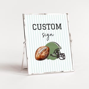 Può includere: Un cartello incorniciato con sfondo a righe verdi e bianche. Il cartello recita "CUSTOM sign" in testo nero. Un pallone da football marrone e un casco da football verde sono illustrati sul cartello.