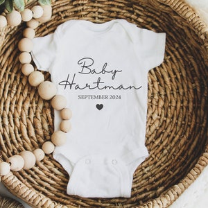May include: White baby onesie with black text that reads "Baby Hartman SEPTEMBER 2024" and a small black heart.