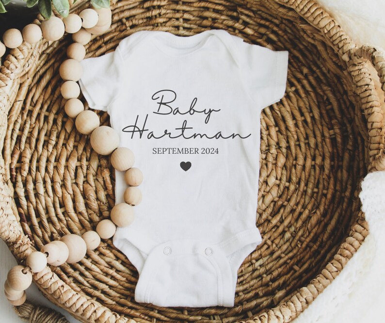 May include: White baby onesie with black text that reads "Baby Hartman SEPTEMBER 2024" and a small black heart.