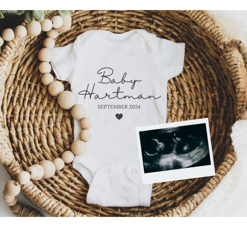 May include: White baby onesie with black text that reads "Baby Hartman SEPTEMBER 2024" and a small black heart. A black and white ultrasound image is next to the onesie.