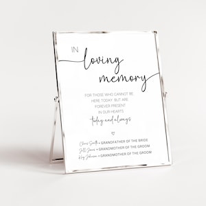 May include: A white sign with black text that reads "In loving memory" and "For those who cannot be here today but are forever present in our hearts today and always." The sign also lists the names of three people and their relationship to the bride and groom.