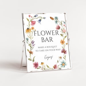 以下が含まれることがあります： 花柄の縁取りが施された白い看板に、黒い文字で「FLOWER BAR」と書かれています。看板には、「持ち帰り用のブーケを作ってください。楽しんで！」とも書かれています。