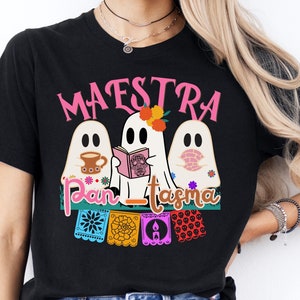May include: Black t-shirt with a pink "MAESTRA" text and three ghosts wearing Day of the Dead masks. The ghosts are holding a book, a cup of coffee, and a flower. The text "Pan-tasma" is below the ghosts. The shirt has a colorful Day of the Dead design.