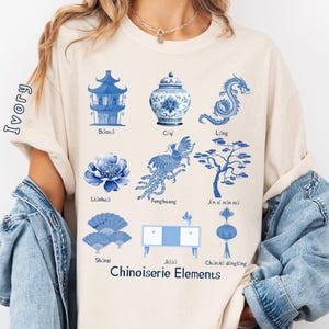 Puede incluir: Una camiseta beige con un estampado azul y blanco de elementos de diseño chino. El estampado incluye una pagoda, un jarrón, un dragón, un fénix, un árbol, un abanico, una mesa y una linterna. El texto "Chinoiserie Elements" está impreso debajo del diseño.