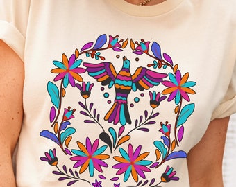 May include: A beige t-shirt with a colorful floral design featuring a bird in the center. The design is reminiscent of Mexican folk art.