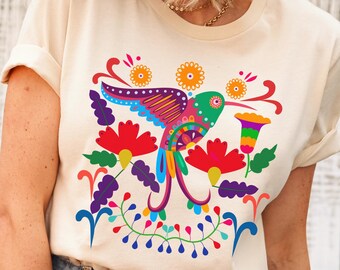 May include: A cream colored t-shirt with a colorful hummingbird and floral design. The hummingbird is pink, green, blue, and yellow. The flowers are red, yellow, and purple.