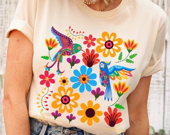 May include: A cream colored t-shirt with a colorful floral and hummingbird design. The design features two hummingbirds, one blue and one green, flying among pink, yellow, orange, and green flowers.