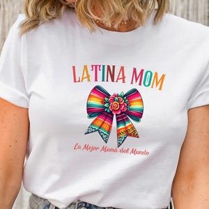 Puede incluir: Camiseta blanca con el texto "LATINA MOM" en letras coloridas. Un diseño de lazo vibrante con una flor está debajo del texto, y la frase "La Mejor Mamá del Mundo" está impresa debajo del lazo.