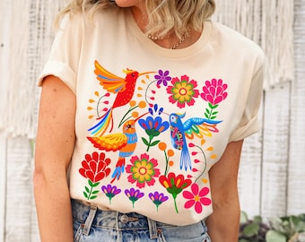 May include: A beige t-shirt with a colorful floral and hummingbird design. The design features three hummingbirds with bright red, yellow, and blue feathers, surrounded by flowers in shades of pink, red, yellow, and purple.